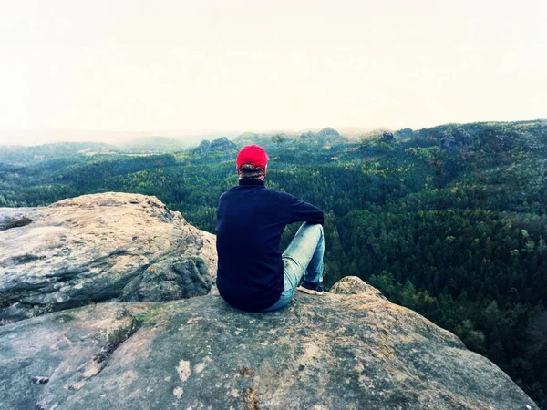 Wanderer auf Klippe. Tourist in roter Mütze, schwarzem Sweatshirt und Jeans sitzt auf scharfer Bergklippe über dem Tal — Stockfoto