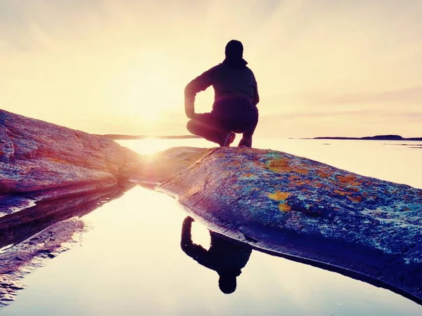 El hombre se sienta al atardecer. Caminante sentarse en cuclillas en acantilado de granito por encima del agua lisa . — Foto de Stock