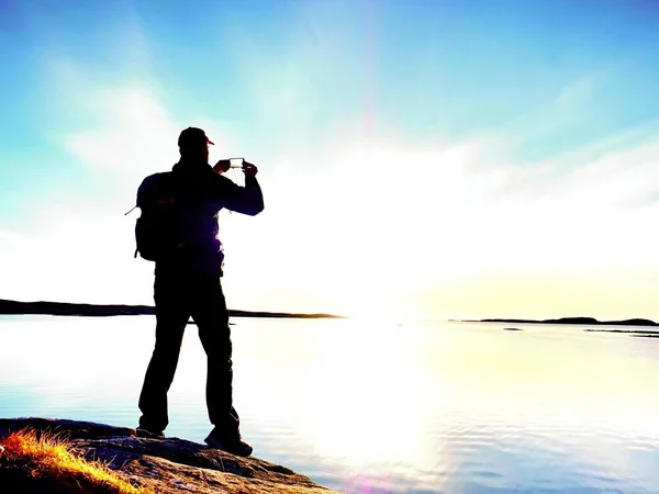 El hombre con chaqueta impermeable en la costa rocosa toma una foto por teléfono inteligente. Fin del maravilloso día — Foto de Stock