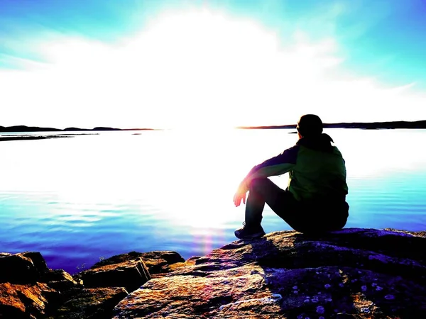 Tired man sit on rocky sea coast.  Calm water, rocky  island and sunset sky background. — Stock Photo, Image