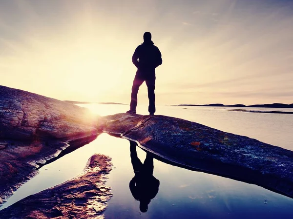 Tall hiker in dark sportswear with backpack stands on cliff above sea. Man enjoy amazing sunset — Stock Photo, Image