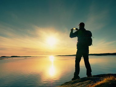 Kayalık sahil şeridi üzerinde su geçirmez ceketli adamı fotoğraf tarafından akıllı telefon alır. Harika gün sonu