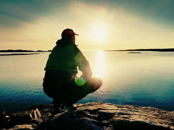 Müder Mann sitzt an felsiger Meeresküste. ruhiges Wasser, felsige Insel und Sonnenuntergangshimmel. — Stockfoto