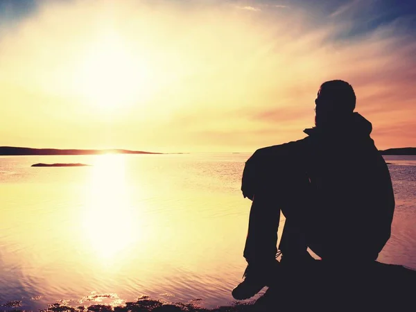 Um homem cansado senta-se na costa rochosa. Água calma, ilha rochosa e pôr do sol céu fundo . — Fotografia de Stock