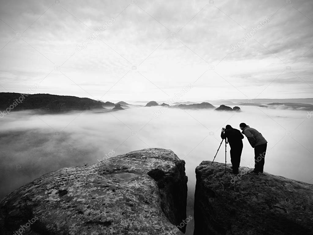 Two warm coat photographers with tripod shoot pictures of autumnal landscape below mountain.