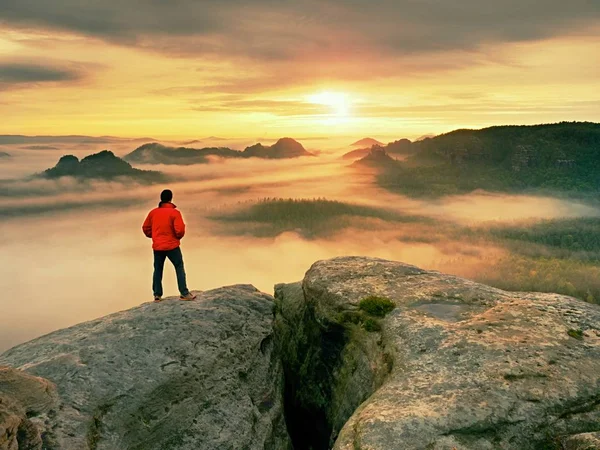 Wanderer in Schwarz auf dem felsigen Gipfel. wunderschöner Tagesanbruch in den Bergen, schwerer orangefarbener Nebel im tiefen Tal. — Stockfoto