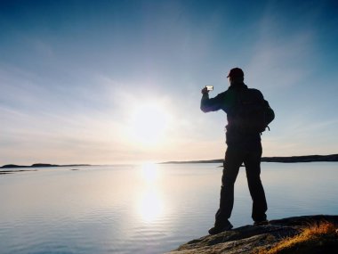 Kayalık sahil şeridi üzerinde su geçirmez ceketli adamı fotoğraf tarafından akıllı telefon alır. Harika gün sonu