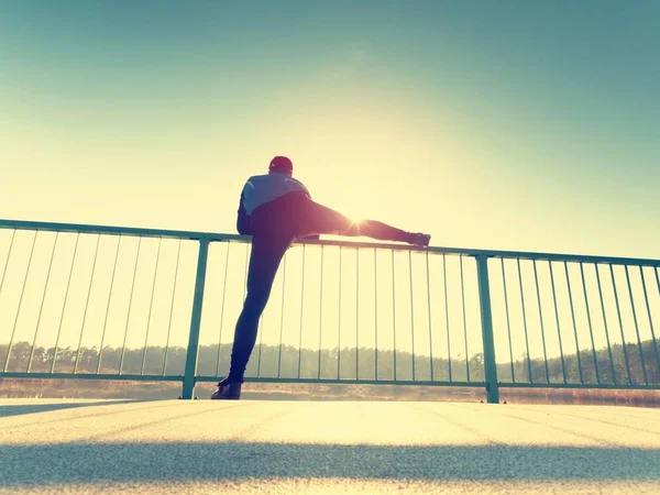 Morning runner in tall black leggings makes body stretching on bridge path. Outdoor exercising