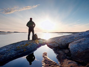 Sırt çantası olan genç duran adam. Uzun yürüyüşe çıkan kimse, renkli gökyüzü günbatımı sahil üzerinde taş.