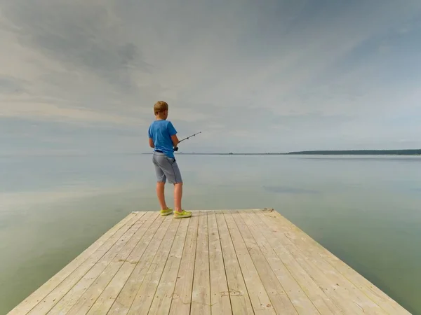 Kleine blond haar jongen is de visserij aan het einde van houten mol. Soepele waterniveau in de baai — Stockfoto