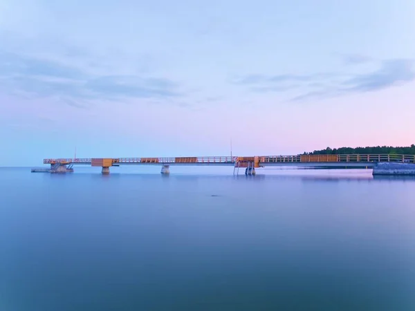 Ponte marítima longa na costa durante a manhã. Tempo frio com primeiros raios de sol no horizonte, água lisa. — Fotografia de Stock