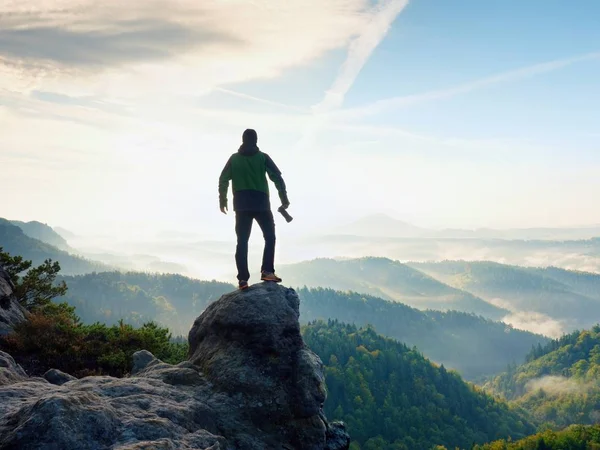 Fotógrafo con cámara en las manos en la cima de la montaña. Caminante escaló en el pico de roca por encima del valle brumoso —  Fotos de Stock