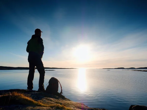 Junger stehender Mann mit Rucksack. Wanderer auf dem Stein am Meeresufer bei buntem Sonnenuntergang. — Stockfoto