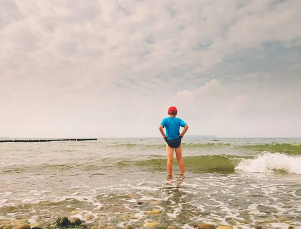 El chico rubio se queda con la marea fría. Niño en la playa de piedra con olas espumosas. Día ventoso —  Fotos de Stock