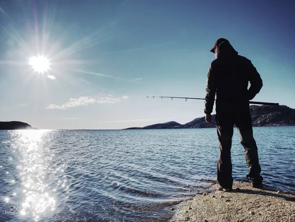 A fiatalember a halászat a tavon naplementekor. Egy horgászbot ember egyedül áll, sziklás partján — Stock Fotó