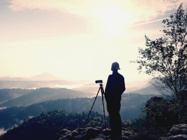 Nature photographer stay at tripod on summit and thinking. Hilly foggy landscape clipart