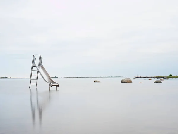 Sunset shore and old steel slide in lake. Chrome ladder tower with  sliding track, big granite stone — Stock Photo, Image