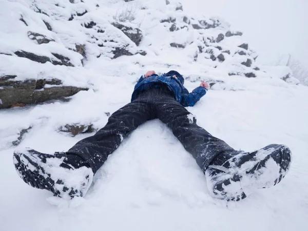 Chico gracioso tirado en la nieve. Juego de niños en nieve fresca en la colina pedregosa —  Fotos de Stock