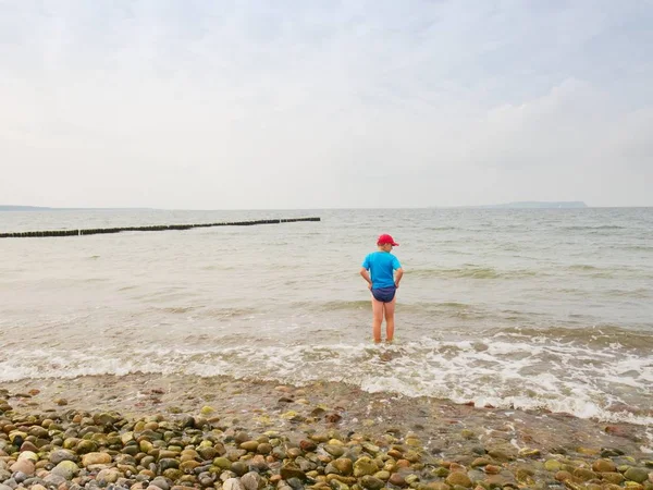 Niño en azul ropa deportiva negro permanecer en el frío mar espumoso. Cabello rubio niño en ondas . —  Fotos de Stock