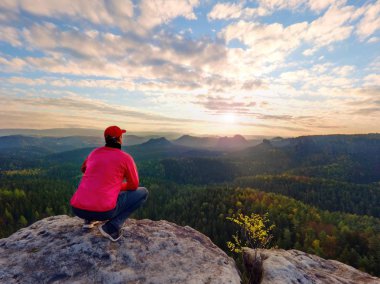 Tek başına kayalıklarda oturan koyu kırmızı açık elbiseler uzun yürüyüşe çıkan kimse arka görünümü. Dağlar vadi, soğuk yukarıda kayalık tepe gizli yağmurlu bulutlar güneş keskin. 
