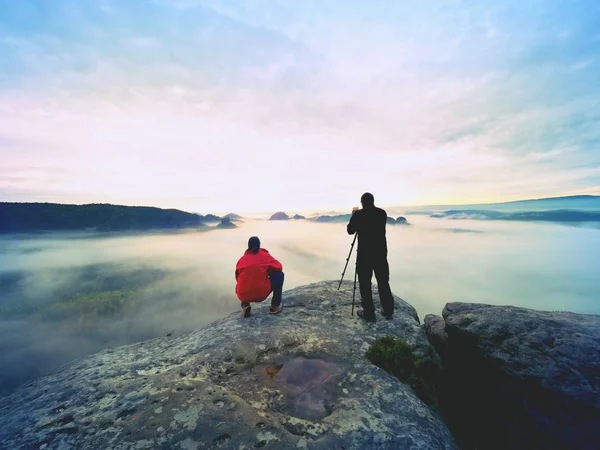 Fotograf mit Auge auf Sucher der Kamera auf Stativ bleibt auf Klippe stehen und macht Fotos, redet mit Freunden. — Stockfoto