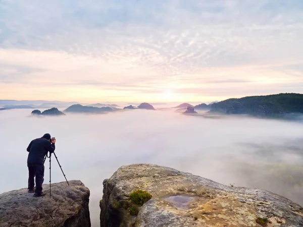 Photographe professionnel au-dessus des nuages. Homme prend des photos avec appareil photo sur trépied sur pic rocheux . — Photo