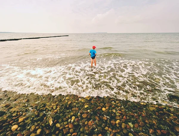 Niño en azul ropa deportiva negro permanecer en el frío mar espumoso. Cabello rubio niño en ondas . —  Fotos de Stock