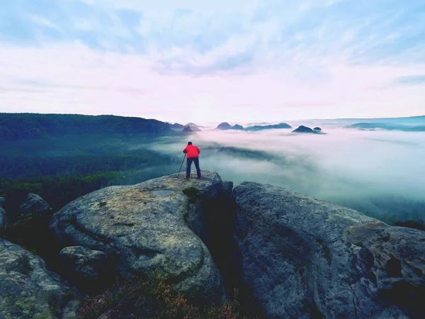 Fotógrafo profissional acima das nuvens. Homem tira fotos com câmera no tripé no pico rochoso . — Fotografia de Stock