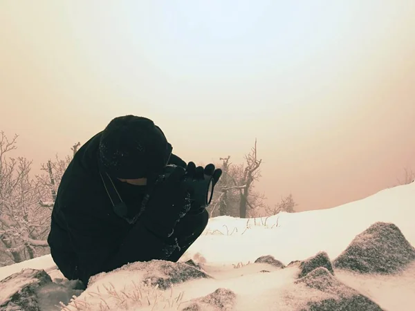 Photographer lay into snow and  taking photo of frozen grass with mirror camera on neck.