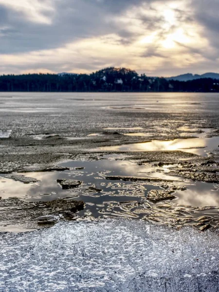 Exposed shore under melting ice. Close up view to border between ice and dark water.