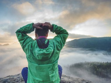Woman sit on cliff edge and looking to rising sun above mist