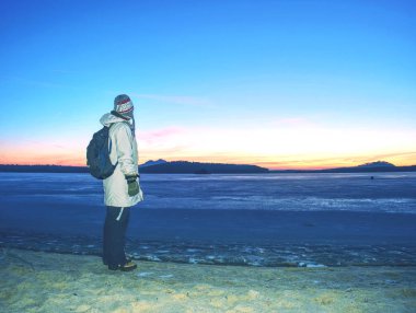 Sıcak uzun ceketli kadın donmuş göl veya nehir de kumlu plajda kalmak. Tepelik ufukta gün tatili ile kış soğuk sabah.
