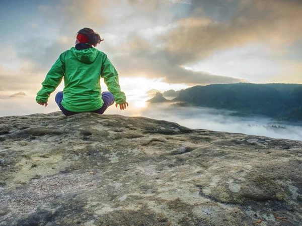 Woman sit on cliff edge and looking to rising sun above mist — Stockfoto