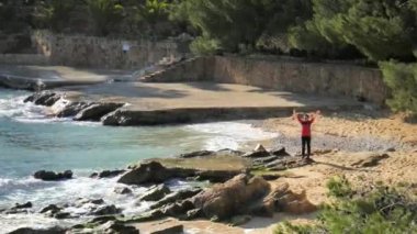 Koyu saçlı kadın deniz taşlı kıyıda yükselmiş kollarıyla selamlamak için el sallıyor. Orta deniz, Mallorka Adası 'nda güneşli bir gün..