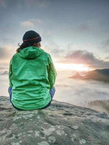 Женщина Зеленой Куртке Задумчиво Сидит Краю Камня Смотрит Туманные Облака — стоковое фото