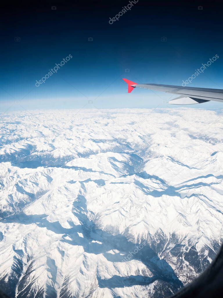 Airplane wing with red winglet float in clear air over snowy winter Swiss Alps.