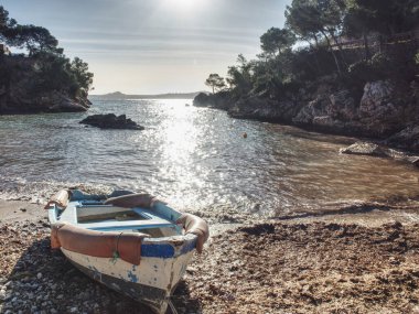 Kayalık koyda terk edilmiş bir balıkçı teknesi güneşe karşı. Yuvarlak koyda özel villası olan kayalık duvarlar. Deniz kıyısındaki kumlu bir sahilde, Mallorca adasında.