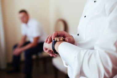 Damat saate bakar ve arka planda bir adam oturur.