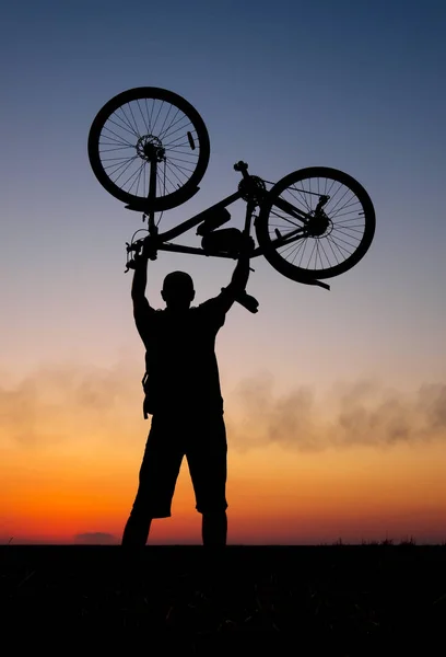 Велосипедист держит свой велосипед над собой на фоне красного заката. Байкер с велосипедом на поле во время восхода солнца — стоковое фото