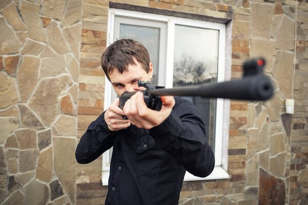 A stern young man with a gun in his hands near the house. 
