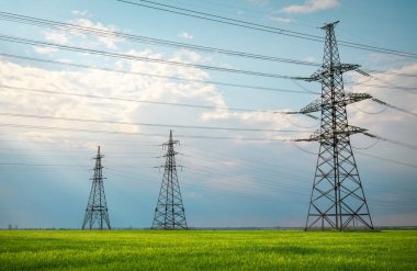 Güneşli bir günde mavi gökyüzünde bulutlar olan düz ve yeşil tarım arazisindeki yüksek voltaj hatları ve elektrik direkleri. Bulutlu ve yağmurlu. Buğday büyüyor.