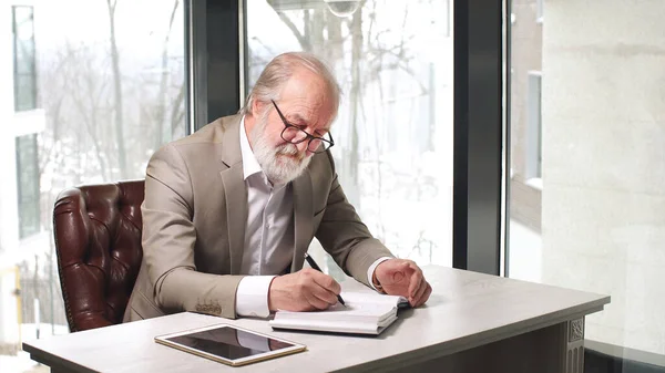 An elderly businessman with a gray beard and gray hair in a classic suit and glasses works in his office