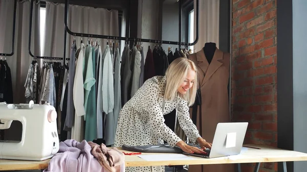 Blonde designer, fashion designer, seamstress smiles while working on a laptop in a clothing workshop