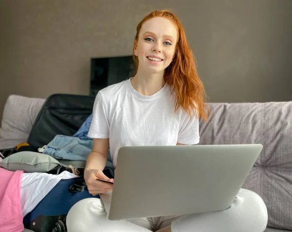 Happy red-haired young woman with a payment card in her hand is sitting next to a suitcase at a laptop. The girl makes purchases over the Internet, buys a ticket, a vacation ticket