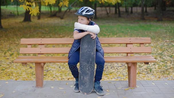 Белый парень со сломанной рукой сидит со скейтом — стоковое видео