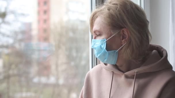 Droevige vrouw met medisch masker bij het raam — Stockvideo