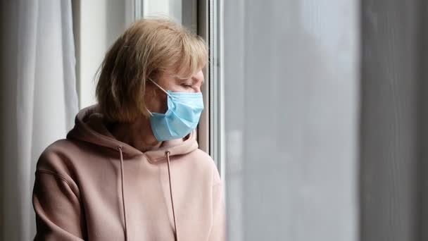 Droevige oudere vrouw met medisch masker bij het raam — Stockvideo