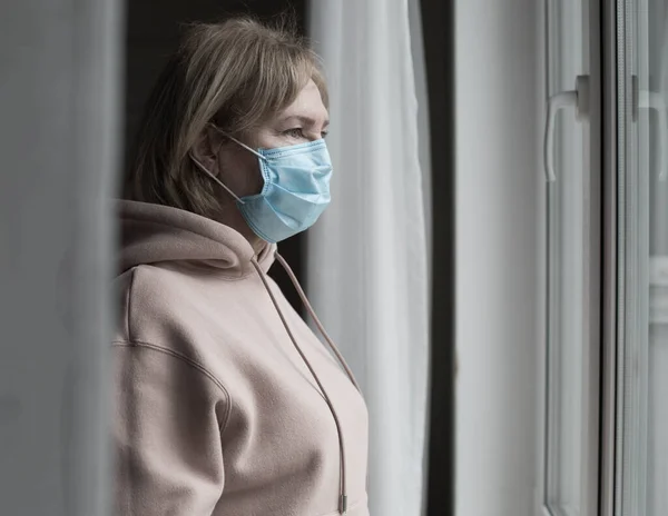 창문 가까이에 의료용 마스크를 쓴 연로 한 여자의 모습 — 스톡 사진