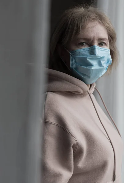 창문 가까이에 의료용 마스크를 쓴 연로 한 여자의 모습 — 스톡 사진