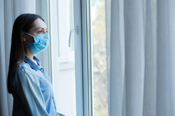 一个戴着医疗面罩的年轻高加索女人站在窗外观看的画像 自我孤立概念 — 图库照片